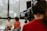 A professional camera operator wearing headphones is filming two people who are sitting in a well-lit room with large windows. The camera display shows the two individuals slightly blurred in the background.