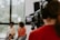 A professional camera operator wearing headphones is filming two people who are sitting in a well-lit room with large windows. The camera display shows the two individuals slightly blurred in the background.