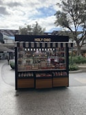 A kiosk with a striped awning displaying a variety of colorful confections and baked goods. The sign above reads 'HOLY CHIC.' The setup is outside, surrounded by trees and other retail structures.