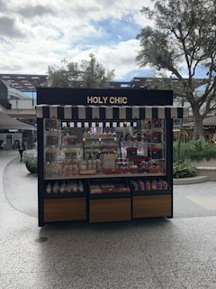 A kiosk with a striped awning displaying a variety of colorful confections and baked goods. The sign above reads 'HOLY CHIC.' The setup is outside, surrounded by trees and other retail structures.
