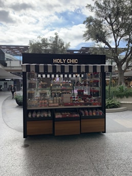 A kiosk with a striped awning displaying a variety of colorful confections and baked goods. The sign above reads 'HOLY CHIC.' The setup is outside, surrounded by trees and other retail structures.