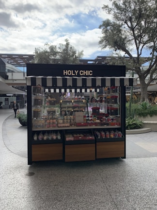 A kiosk with a striped awning displaying a variety of colorful confections and baked goods. The sign above reads 'HOLY CHIC.' The setup is outside, surrounded by trees and other retail structures.