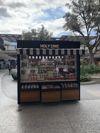 A kiosk with a striped awning displaying a variety of colorful confections and baked goods. The sign above reads 'HOLY CHIC.' The setup is outside, surrounded by trees and other retail structures.