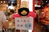 A mannequin head with a black face mask featuring a cartoon bee is displayed inside a shop. The head is mounted on a red stand, and beneath it hangs a sign with Chinese characters, suggesting the importance of wearing a mask. In the background, there are colorful displays and people engaging with merchandise, giving a sense of a busy indoor market or shop.