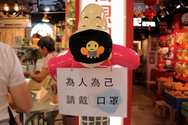 A mannequin head with a black face mask featuring a cartoon bee is displayed inside a shop. The head is mounted on a red stand, and beneath it hangs a sign with Chinese characters, suggesting the importance of wearing a mask. In the background, there are colorful displays and people engaging with merchandise, giving a sense of a busy indoor market or shop.