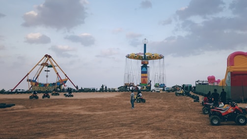 An amusement park setting with a large pirate ship ride on the left and a colorful swing ride in the center. There are several people and children riding small ATVs on the sandy ground, and an inflatable bounce house is visible on the right. The sky is overcast with scattered clouds.