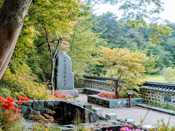 A serene garden scene featuring a stone monument with engraved text, surrounded by vibrant flowers and lush greenery. A stone wall runs parallel to the monument, creating a tranquil atmosphere. The trees and plants display various shades of green, and the overall setting exudes a peaceful, natural ambiance.