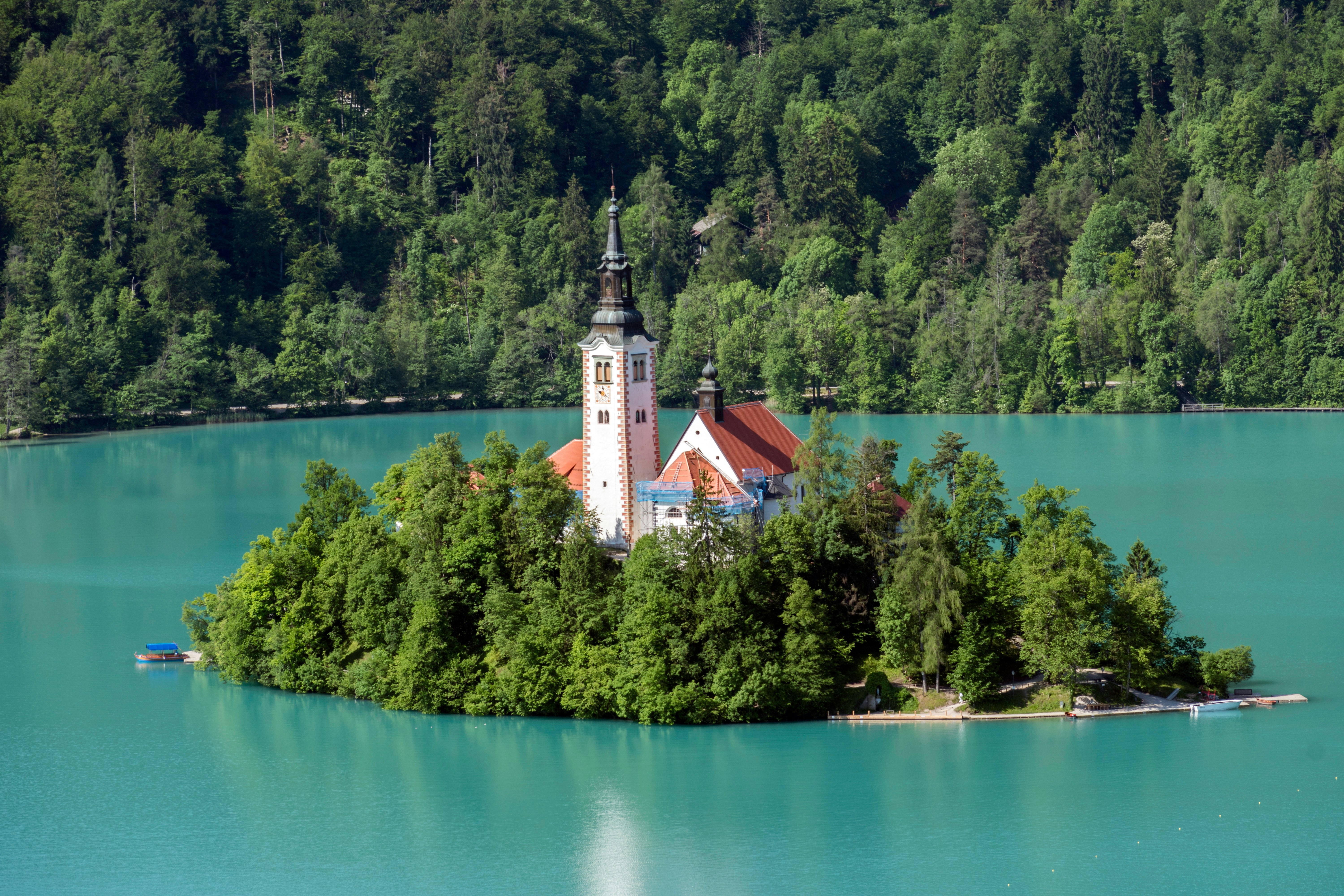 white and brown concrete building near green trees and lake during daytime, A view of Lake Bled and within the middle of the lake is Slovenia’s only natural island. The island has several buildings, the main one being the Pilgrimage Church of the Assumption of Mary, built in the 15th century.