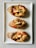 Three baked bread rolls topped with melted cheese and a drizzle of tomato sauce are neatly arranged on a rectangular white ceramic plate. Each roll has a crispy, golden-brown crust with a slightly charred appearance, and the cheese is slightly golden and oozing over the edges.