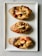 Three baked bread rolls topped with melted cheese and a drizzle of tomato sauce are neatly arranged on a rectangular white ceramic plate. Each roll has a crispy, golden-brown crust with a slightly charred appearance, and the cheese is slightly golden and oozing over the edges.
