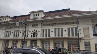A historic European-style building with a white facade featuring large windows and decorative elements. The building has a red-tiled roof and contains signage in Dutch. There are several people on motorcycles and a car in front of the building.