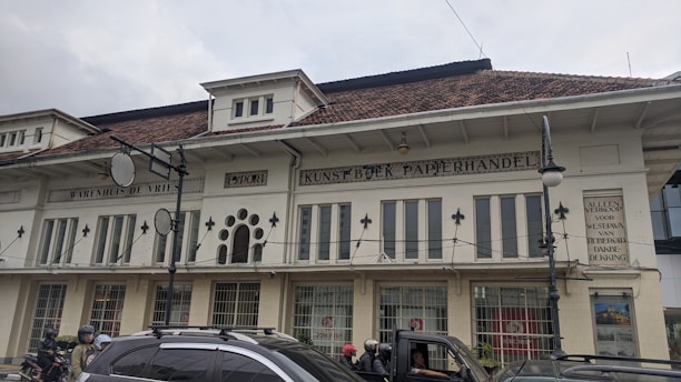 A historic European-style building with a white facade featuring large windows and decorative elements. The building has a red-tiled roof and contains signage in Dutch. There are several people on motorcycles and a car in front of the building.