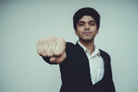A person dressed in a black suit and white shirt is extending their fist toward the camera, creating a dynamic and assertive pose. The background is plain and soft in color, adding focus to the subject.