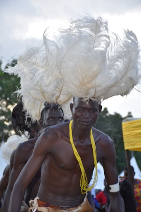 Several people are participating in a cultural dance or ceremony, wearing elaborate headgear made of white feathers. One person is prominently in the foreground and is adorned with a yellow necklace and a white armband. The background suggests an outdoor setting with trees and some colorful structures.