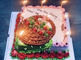 A colorful birthday cake adorned with lit candles on top. The cake features elaborate decorations, including red and green cherries, kiwi slices, and chocolate icing. Floral decorations in pink, red, and green are noticeable along the sides and top borders of the cake.