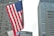 An American flag waves prominently in the foreground against a backdrop of tall skyscrapers, including a building with the MetLife logo visible. The image conveys a sense of urban environment with a focus on national pride.