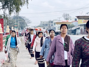 A group of women walking on a street while balancing trays of items on their heads. They are wearing a mix of traditional and modern clothing with long skirts and jackets. The background includes urban elements such as buildings, vehicles, and power lines, along with a few trees.