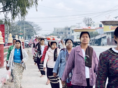 A group of women walking on a street while balancing trays of items on their heads. They are wearing a mix of traditional and modern clothing with long skirts and jackets. The background includes urban elements such as buildings, vehicles, and power lines, along with a few trees.