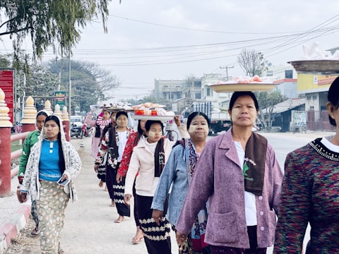 A group of women walking on a street while balancing trays of items on their heads. They are wearing a mix of traditional and modern clothing with long skirts and jackets. The background includes urban elements such as buildings, vehicles, and power lines, along with a few trees.