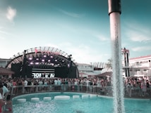 A large outdoor music event taking place in front of a stage with bright lights and a prominent sign above. The crowd is gathered around a swimming pool with water features and circular seats. People are enjoying the vibrant atmosphere, with modern architecture and palm trees in the background.