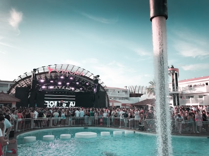 A large outdoor music event taking place in front of a stage with bright lights and a prominent sign above. The crowd is gathered around a swimming pool with water features and circular seats. People are enjoying the vibrant atmosphere, with modern architecture and palm trees in the background.