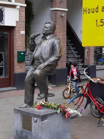 A bronze statue of a man holding a microphone is seated on a pedestal with flowers placed on it. Behind the statue, there is a young child wearing a hat and a red jacket riding a bicycle. The background shows a building with brick and white walls, along with a large yellow sign on the right side. Stairs leading to a doorway can also be seen in the background.