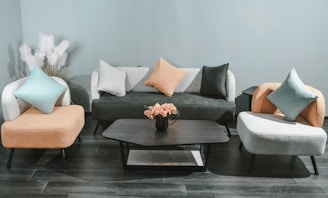 A modern living room setting with a minimalist design features a black sofa adorned with white, black, and orange pillows. Two matching armchairs in contrasting colors flank the sofa. A black coffee table stands in the center, topped with a vase of pink flowers. In the background, decorative white pampas grass adds texture against the light blue wall. The floor is lined with dark wood panels, enhancing the contemporary feel.