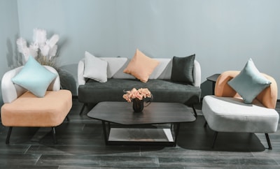A modern living room setting with a minimalist design features a black sofa adorned with white, black, and orange pillows. Two matching armchairs in contrasting colors flank the sofa. A black coffee table stands in the center, topped with a vase of pink flowers. In the background, decorative white pampas grass adds texture against the light blue wall. The floor is lined with dark wood panels, enhancing the contemporary feel.