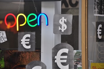 A brightly colored neon sign spells out the word 'open' in red, yellow, green, and blue. Surrounding the sign are printed sheets displaying large currency symbols, including the dollar sign and the euro sign, in monochrome colors. These sheets are affixed to a glass window or door, hinting at a storefront or business setting.