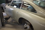 A vintage car is elevated on a car lift in a garage. The vehicle's rear wheel is removed, exposing the brake disc and other components. The car has a sleek, shiny exterior and a classic design. The interior of the garage is dimly lit, with various tools and a covered object in the background.