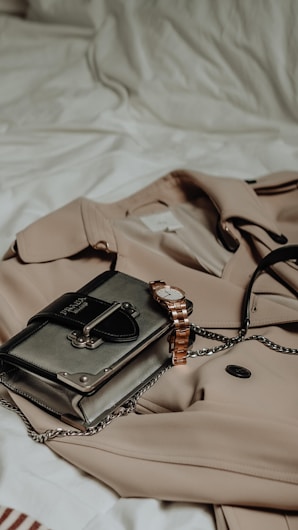 A beige trench coat is laid on a white bed, accompanied by a stylish handbag with metallic accents and a gold wristwatch. The setting suggests a chic and elegant style, with a focus on fashion accessories.