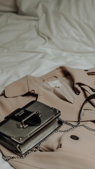A minimalist flat lay of neutral-toned wardrobe essentials including a beige trench coat, white blouse, and classic leather handbag on a clean white background.