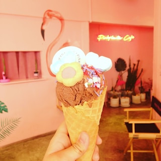 A hand holding a cone of chocolate ice cream, adorned with colorful sprinkles and a cookie, is in focus. The background features a pink-themed interior with potted plants, a flamingo mural, and a neon sign that reads 'Pinkholic Café'.