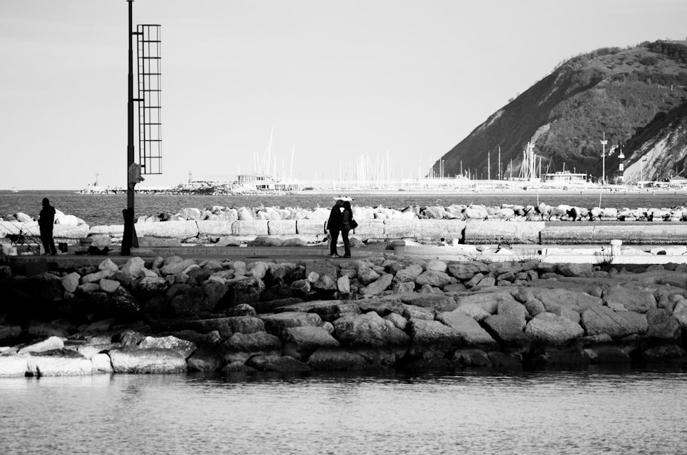 grayscale photo of man and woman standing on rock near body of water