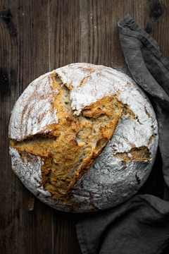 A rustic, round loaf of bread with a cracked, golden-brown crust and a dusting of flour rests on a textured wooden surface. A dark cloth lies beside it, adding to the warm, artisanal feel.
