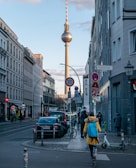 A city street lined with buildings features a prominent television tower in the background. Several pedestrians walk on the sidewalk, and parked cars line the street. Traffic signs and a bicycle are visible, creating an urban atmosphere.