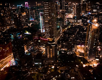 A vibrant cityscape at night with numerous illuminated skyscrapers and buildings. The urban landscape features bright neon lights and tall structures, creating a lively atmosphere. Streets are visible below, with light trails from traffic adding dynamism to the scene.