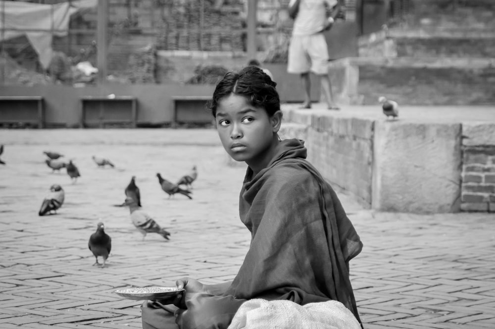 grayscale photo of boy in jacket and pants sitting on concrete floor with birds