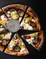 Slices of gourmet pizza topped with roasted potatoes, ricotta cheese, and zucchini are arranged on a dark background. A pizza cutter is placed on one of the slices, enhancing the presentation.
