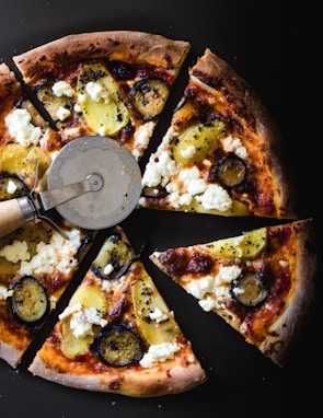 Slices of gourmet pizza topped with roasted potatoes, ricotta cheese, and zucchini are arranged on a dark background. A pizza cutter is placed on one of the slices, enhancing the presentation.
