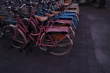 A neatly arranged row of bicycles with colorful frames and baskets, lined up on a paved sidewalk. Each bike has a wooden seat and an advertisement panel on the back, suggesting they might be part of a rental service.