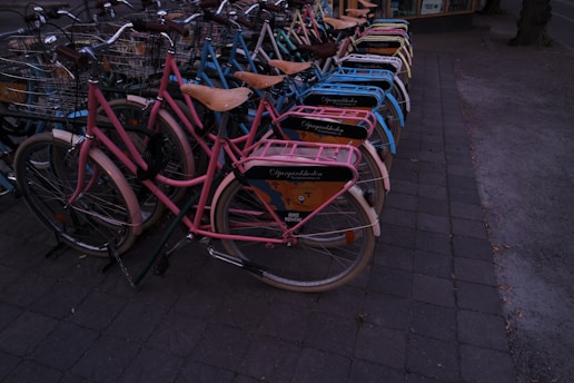A neatly arranged row of bicycles with colorful frames and baskets, lined up on a paved sidewalk. Each bike has a wooden seat and an advertisement panel on the back, suggesting they might be part of a rental service.