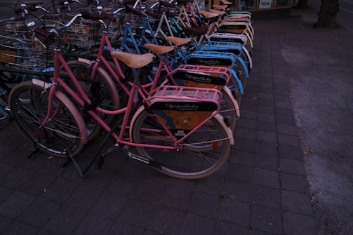 A neatly arranged row of bicycles with colorful frames and baskets, lined up on a paved sidewalk. Each bike has a wooden seat and an advertisement panel on the back, suggesting they might be part of a rental service.
