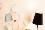A variety of sticky notes with handwritten text are attached to a white wall, displaying what seems to be study materials or reminders. A black desk lamp is positioned nearby, casting light on the notes.