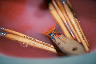 Multiple paintbrushes with wooden handles are submerged in a pinkish liquid, possibly paint or water mixed with paint. One of the brushes has visible blue and orange paint on the bristles.