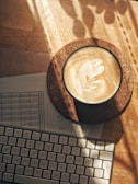 A cup of coffee with latte art placed on a cork coaster is next to a computer keyboard. Sunlight creates shadows across the wooden surface.
