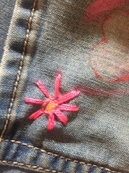 Pink embroidered flower on a denim fabric with visible stitching. The embroidery has a small brown center and several elongated petals.