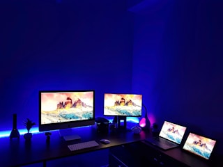 A dark room with illuminated screens set on a desk. Multiple computer monitors and laptops display vibrant images of a mountain landscape. Blue ambient lighting enhances the modern aesthetic of the workspace, with various electronic accessories and decor items like a small plant and decorative lamps contributing to the setup.