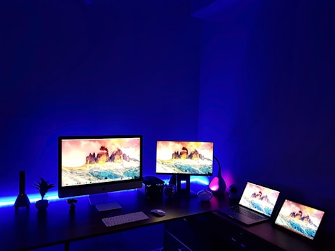 A dark room with illuminated screens set on a desk. Multiple computer monitors and laptops display vibrant images of a mountain landscape. Blue ambient lighting enhances the modern aesthetic of the workspace, with various electronic accessories and decor items like a small plant and decorative lamps contributing to the setup.