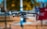 A drone, specifically a Mavic 2, is captured mid-flight with a blurred background of trees and a bright blue sky. The focus on the drone highlights its propellers and camera, suggesting a stable hover. The setting appears sunny, accentuating the metallic surface of the drone.